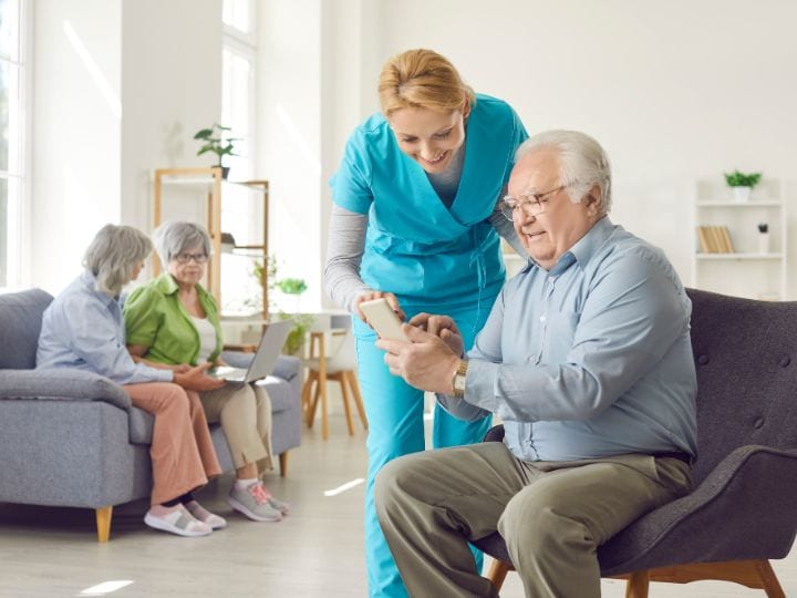 Caregiver in scrubs helping seated elderly man use a smartphone